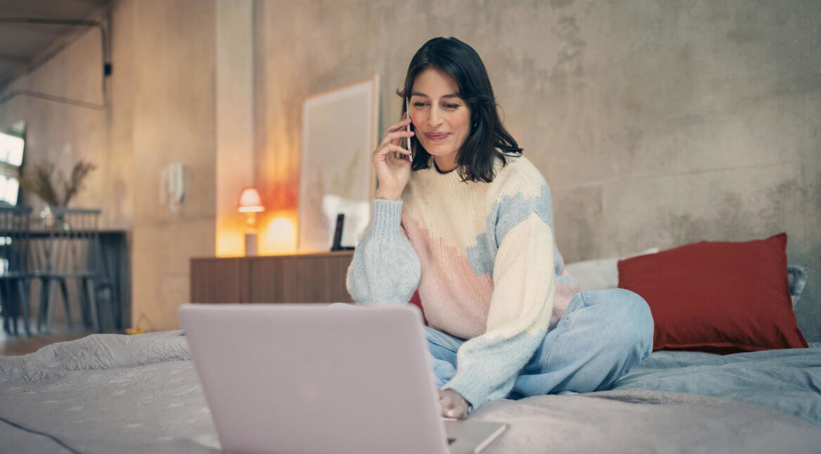 Vodafone Kabel-Internet- & Telefon-Anschluss freischalten: Frau sitzt telefonierend vor ihrem Laptop auf dem Bett.