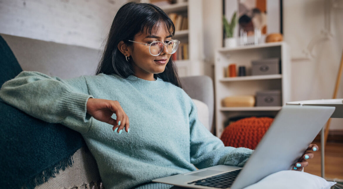 Auf dem Bild sitzt eine junge Frau mit langem dunklem Haar und Brille entspannt auf einem Sofa und arbeitet an einem Laptop. Sie trägt einen hellblauen Pullover und wirkt konzentriert. Das Wohnzimmer im Hintergrund ist modern eingerichtet. Die Szene symbolisiert den alltäglichen Umgang mit ChatGPT-Modelle im privaten Umfeld.