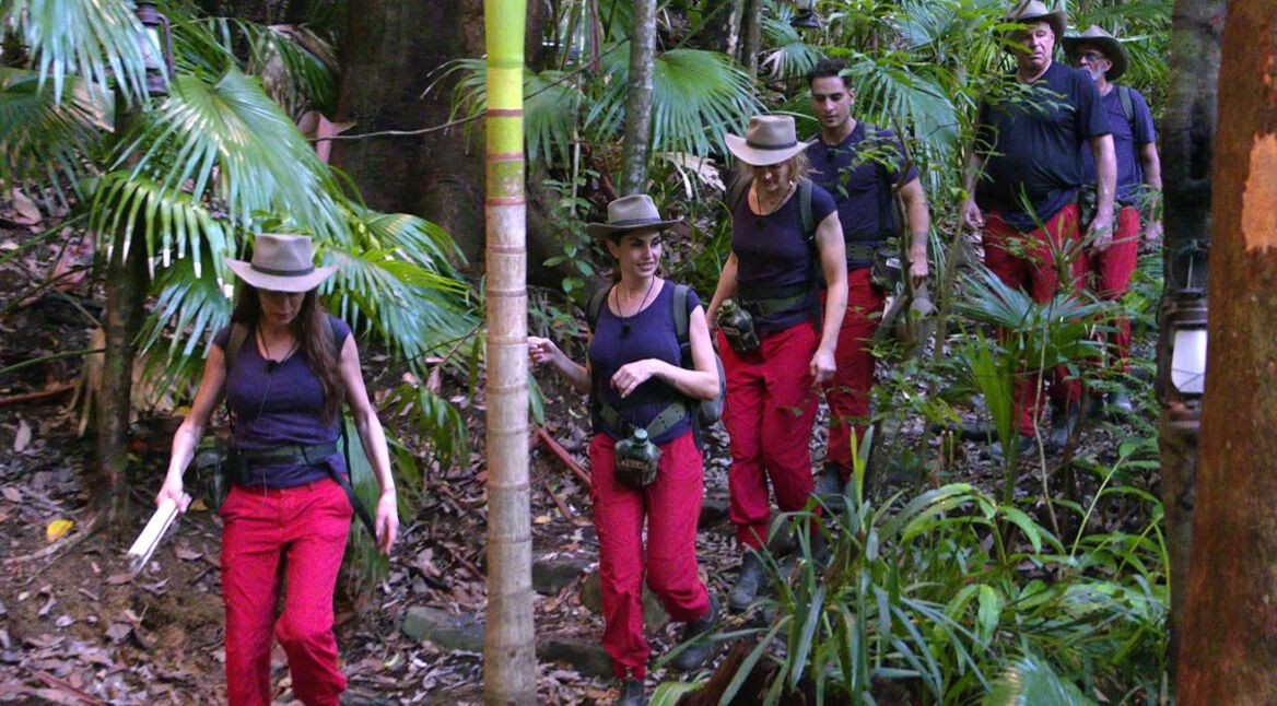 Das Bild zeigt mehrere Kandidat:innen der Staffel 2025 auf dem Weg durch den dichten Urwald. Sie tragen typische Dschungelcamp-Outfits mit roten Hosen, dunklen Shirts und Hüten. Die Gruppe bewegt sich konzentriert durch das grüne, feuchte Dickicht. Das Bild symbolisiert den harten Weg der Dschungelcamp-Gewinner:innen und ihre Herausforderungen im australischen Busch.