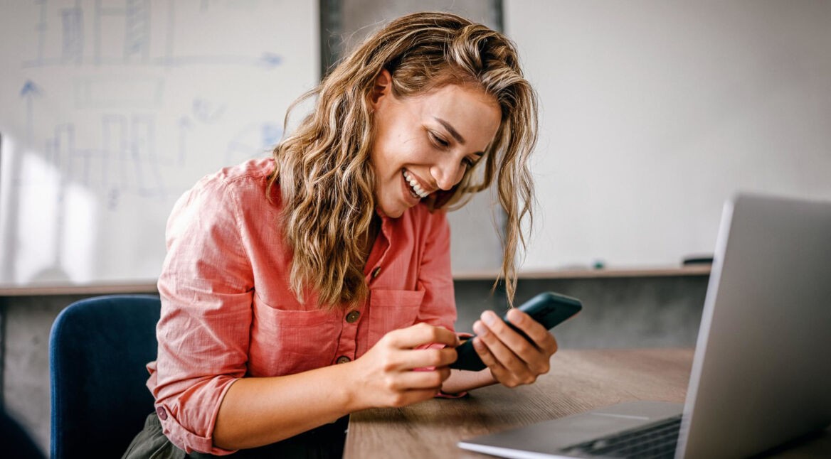 iPhone WLAN Hotspot einrichten: Frau sitzt lachend mit ihrem Smartphone in der Hand vor ihrem Laptop.