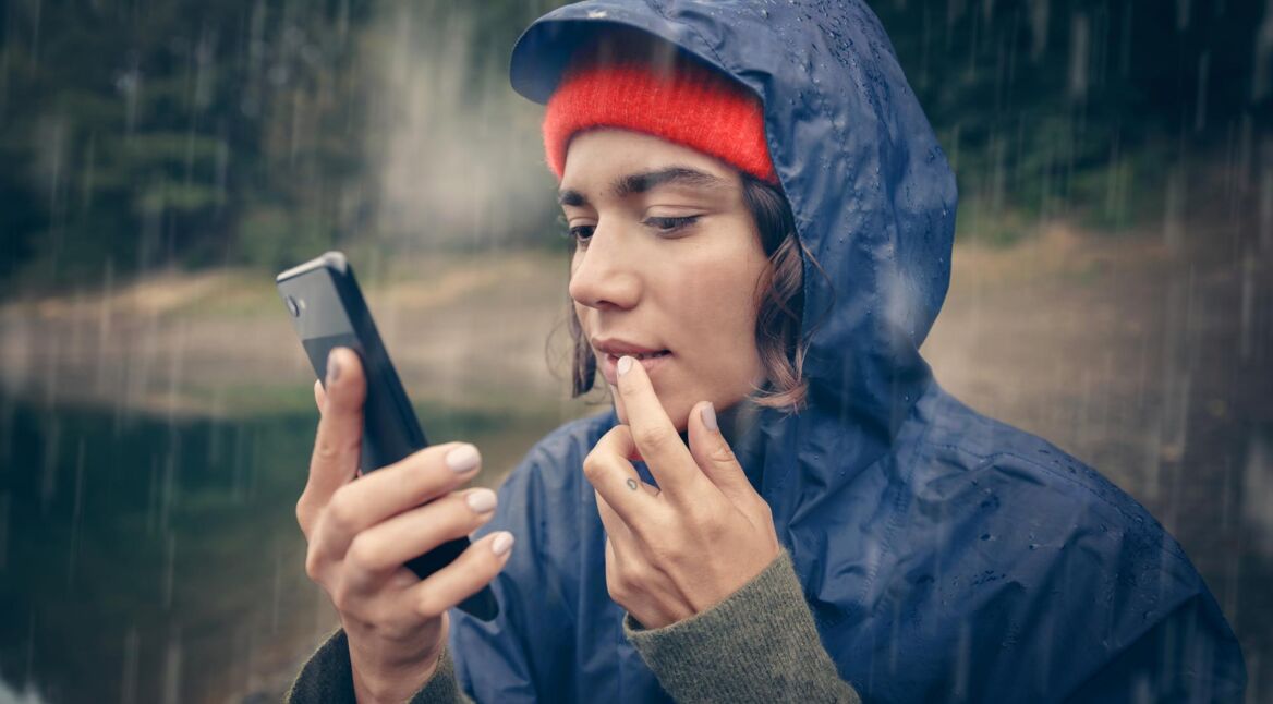 Kein Internet trotz Roaming: Junge Frau in Regemantel benutzt im Regen ihr Handy
