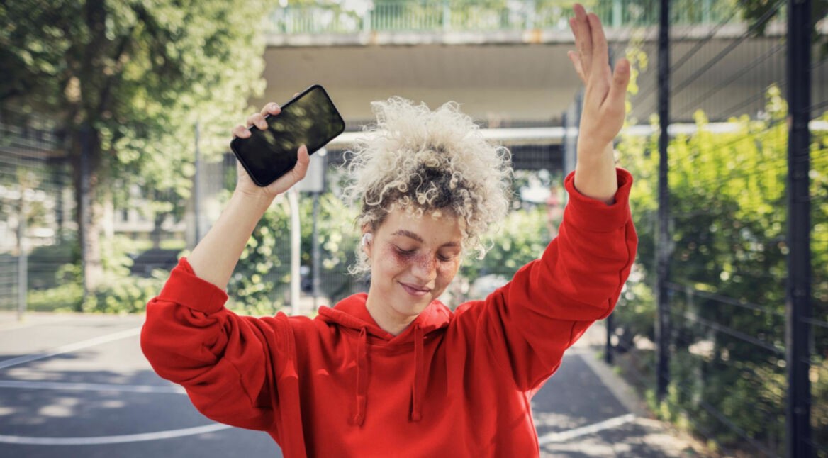 Datenverbrauch Social Media: Junge blonde Frau mit lockigen Haaren tanzt ausgelassen mit ihrem Handy in der Hand.