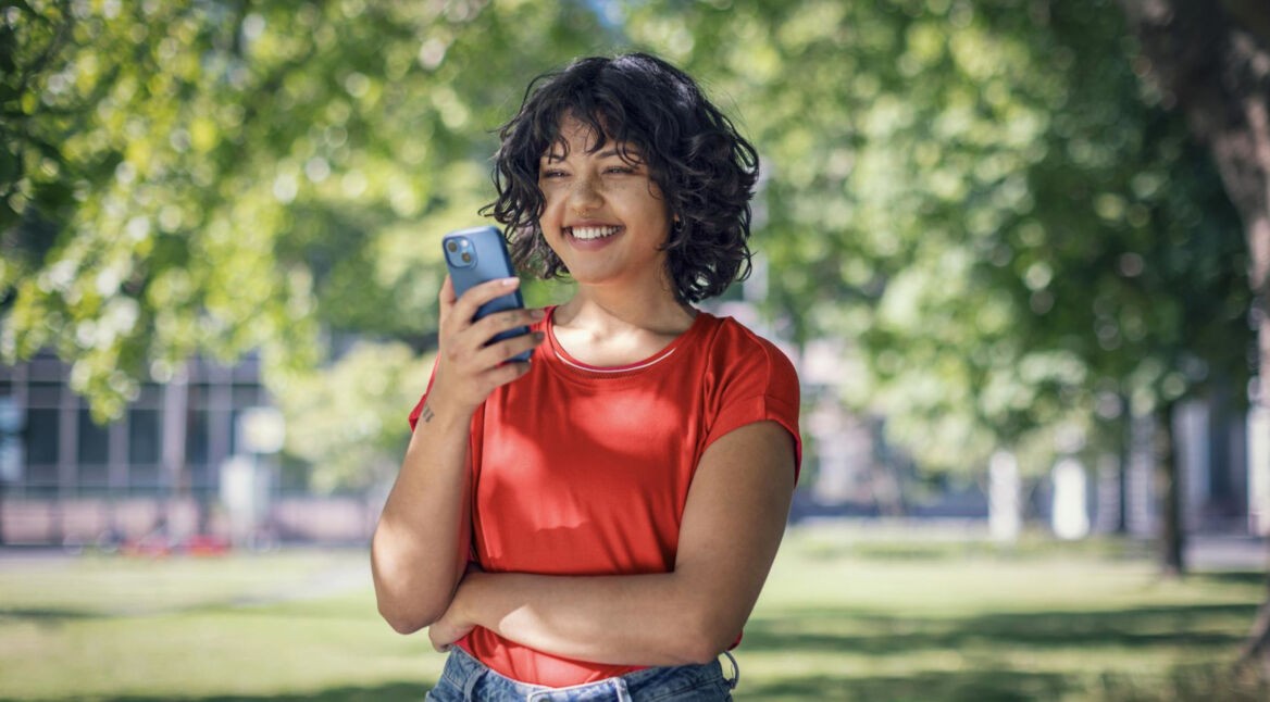 Plötzlich hoher Datenverbrauch: junge Frau schaut lächelnd auf ihr Handy in einem Park.