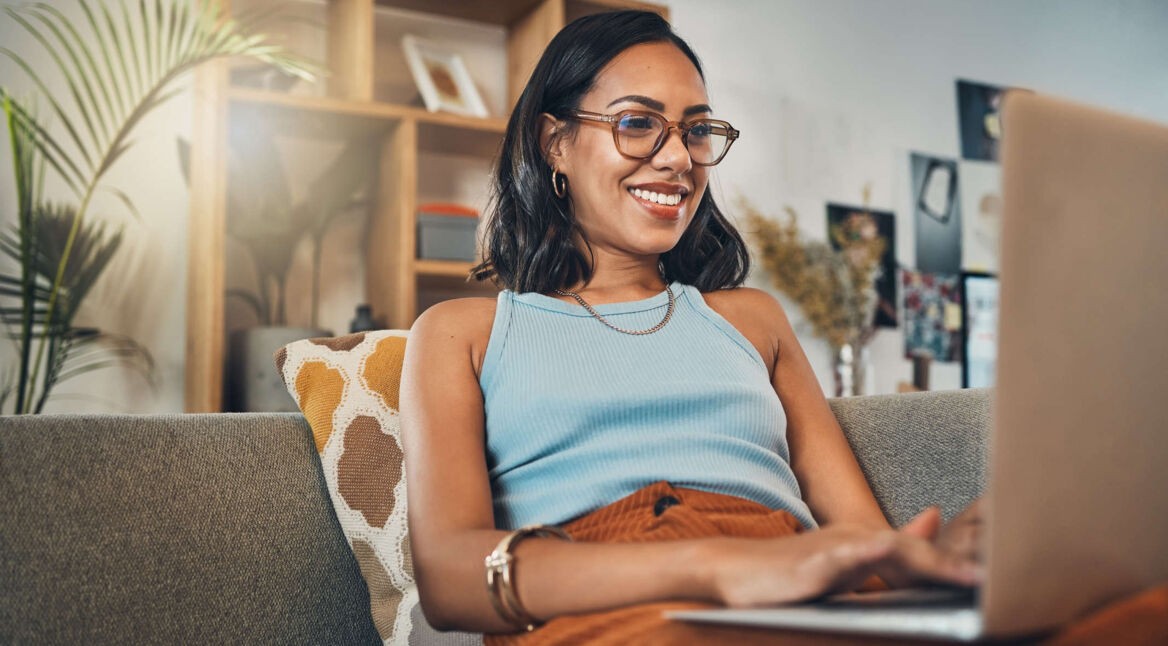 Eine Frau sitzt mit einem Laptop auf dem Schoß auf einer Couch.