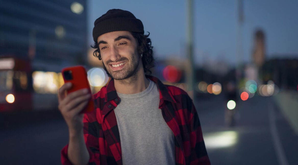 Roaming Android aktivieren: Mann in kariertem Hemd steht abends auf der Straße und schaut lächelnd auf sein Handy