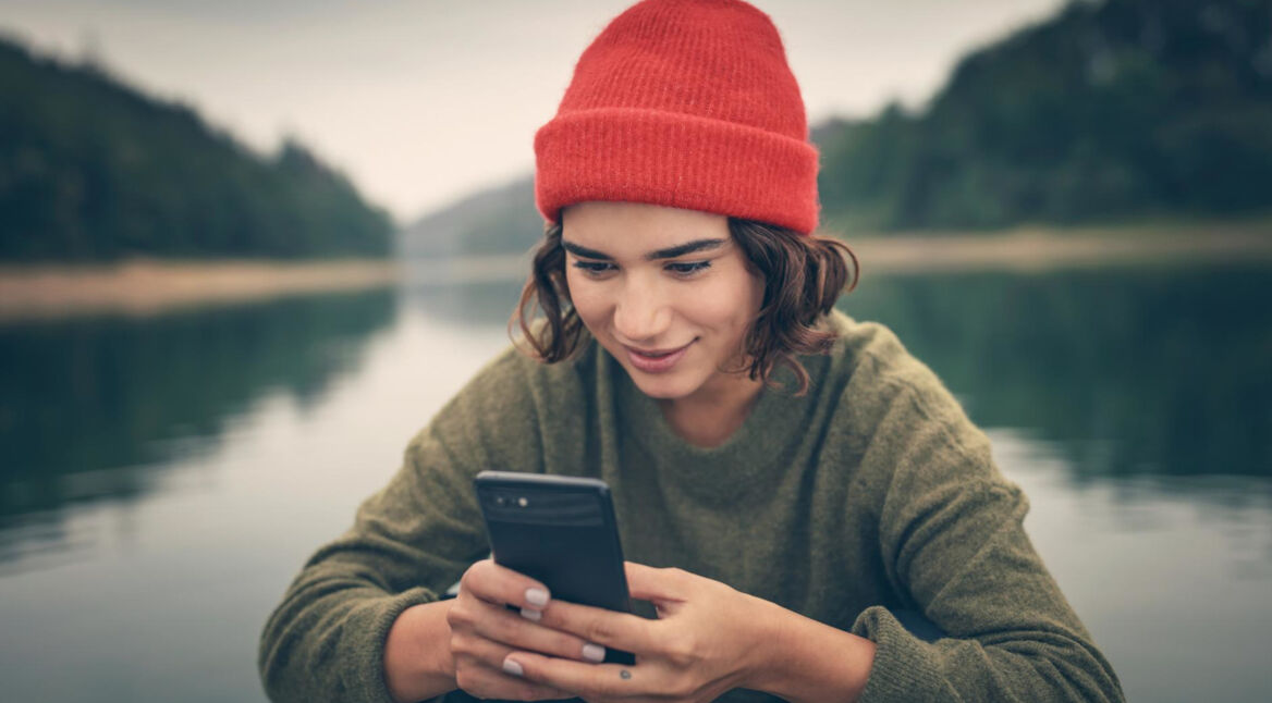 Roaming Samsung: Junge Frau sitzt mit einer roten Mütze und ihrem Handy in der Hand an einem See