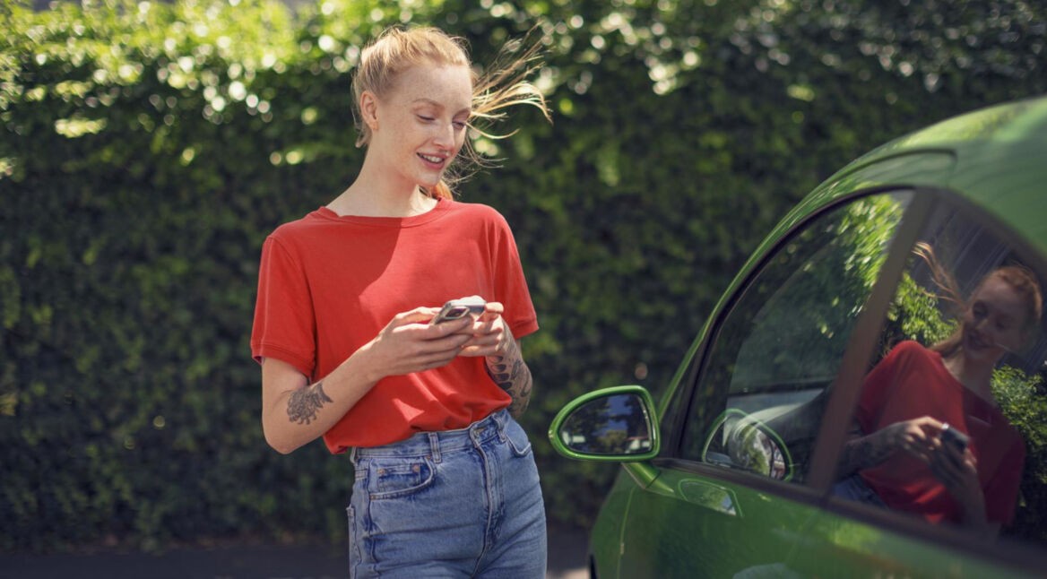Roaming am iPhone aktivieren: Junge Frau mit ihrem Handy läuft an einem grünen Auto vorbei