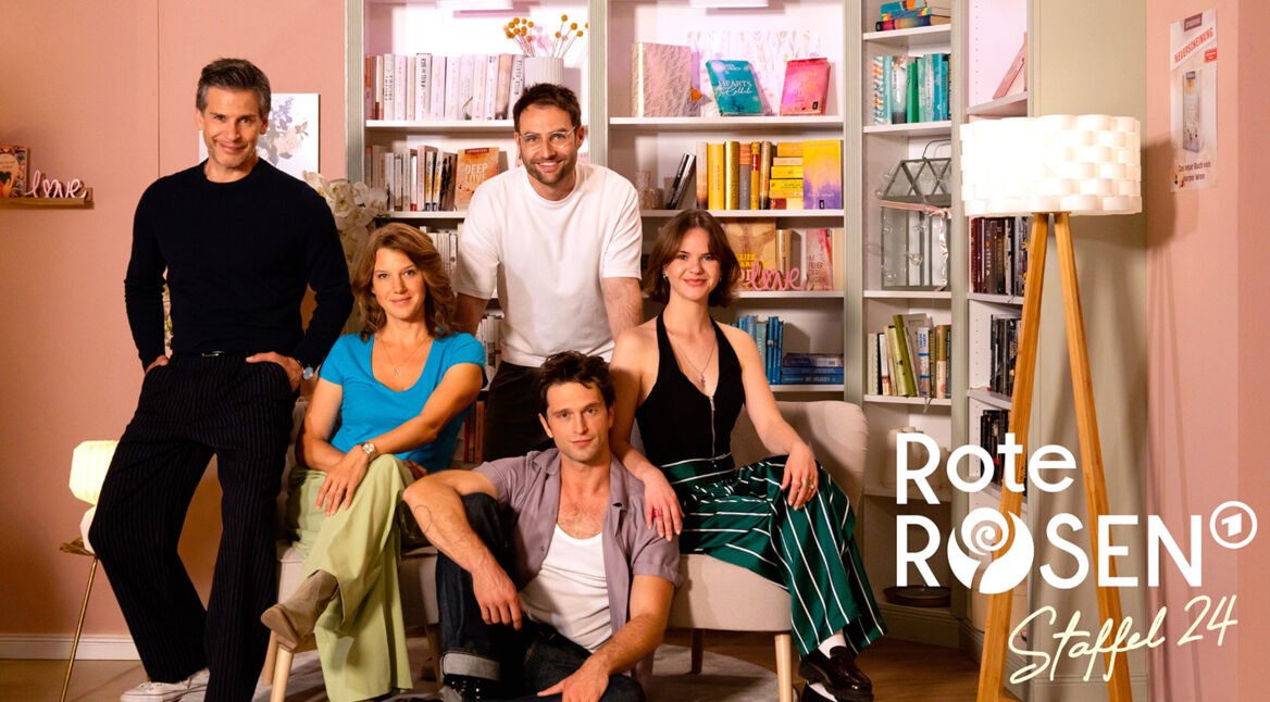 Auf dem Bild zur Rote Rosen Staffel 24 posieren fünf Schauspieler:innen vor einem hellen Bücherregal. Zwei Personen sitzen auf einem Sofa, drei stehen oder sitzen locker drumherum.