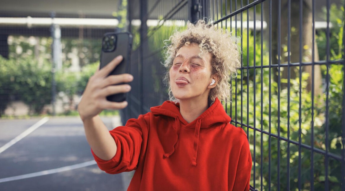 WhatsApp Datenverbrauch: Junge Frau steht draußer in urbaner Umgebung und streckt die Zunge in Richtung ihrer Handykamera heraus.