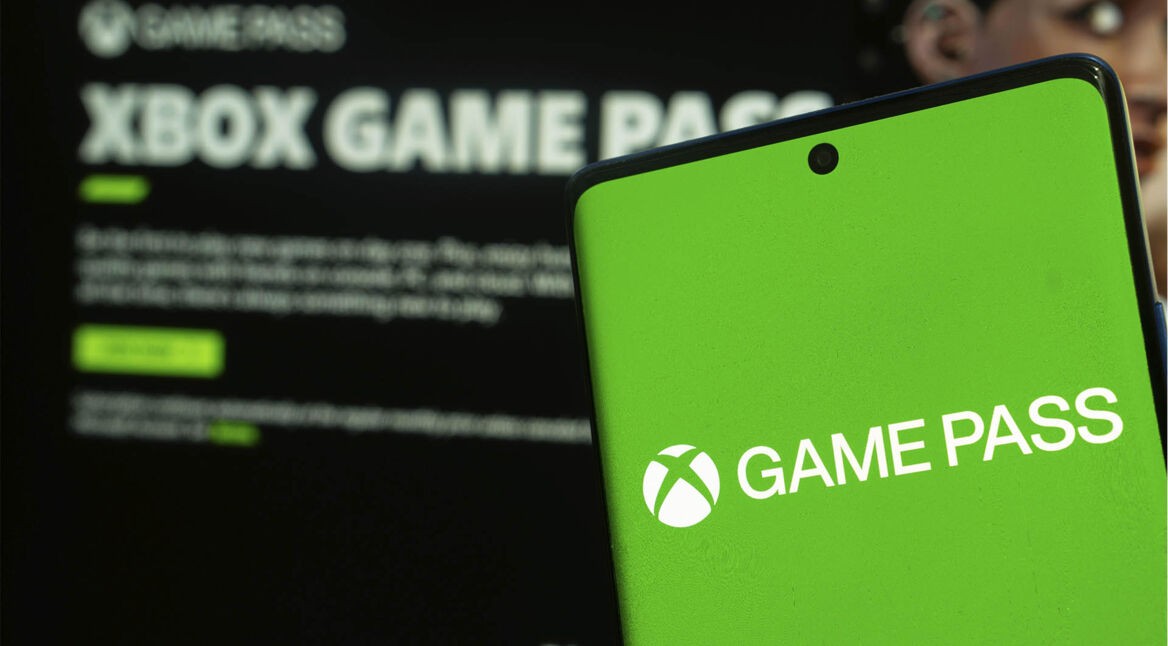 Ein Smartphone zeigt das Logo des Xbox Game Pass; im Hintergrund ist die geöffnete Game-Pass-App unscharf zu erkennen.
