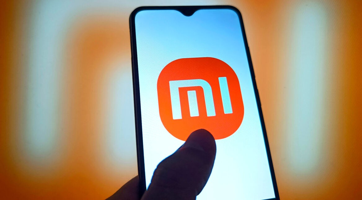 Smartphone mit Mi-Logo und einem Daumen auf dem Display vor orangefarbenem Hintergrund