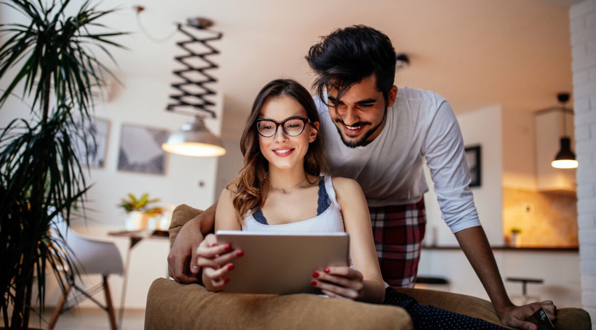Ein junges Paar schaut gemeinsam auf ein Tablet und lächelt dabei.