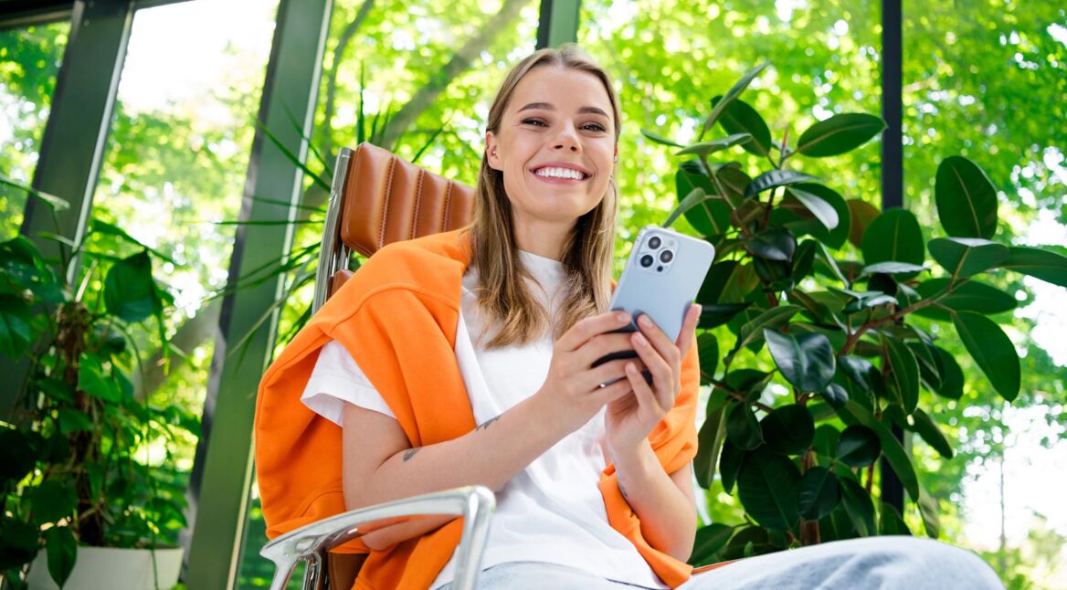 Auf dem Bild in dem Artikel zur Samsung Garantie sitzt eine junge Frau entspannt in einem modernen, hellen Raum mit vielen Pflanzen und hält ein Smartphone in der Hand.