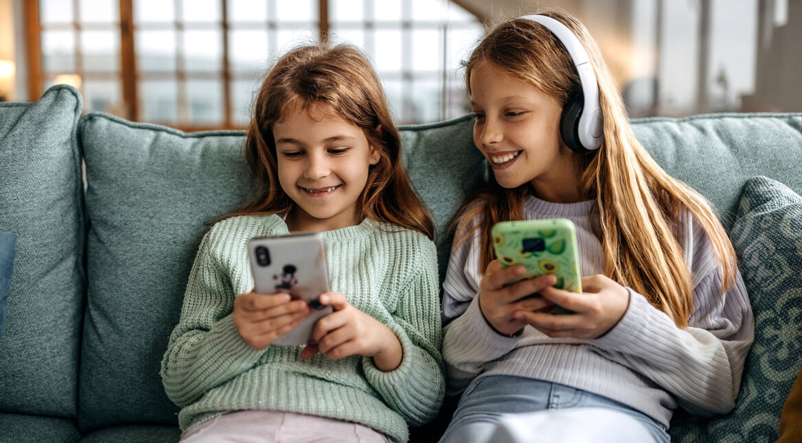 Auf dem Bild zum Thema Smartphones für Kinder sitzen zwei Mädchen fröhlich nebeneinander auf einem Sofa. Beide halten jeweils ein Smartphone in der Hand, eines mit bunter Hülle, das andere mit Tiermotiv. Das Mädchen rechts trägt zusätzlich große weiße Kopfhörer und lächelt ihre Freundin an. Die Szene wirkt entspannt und spielerisch.
