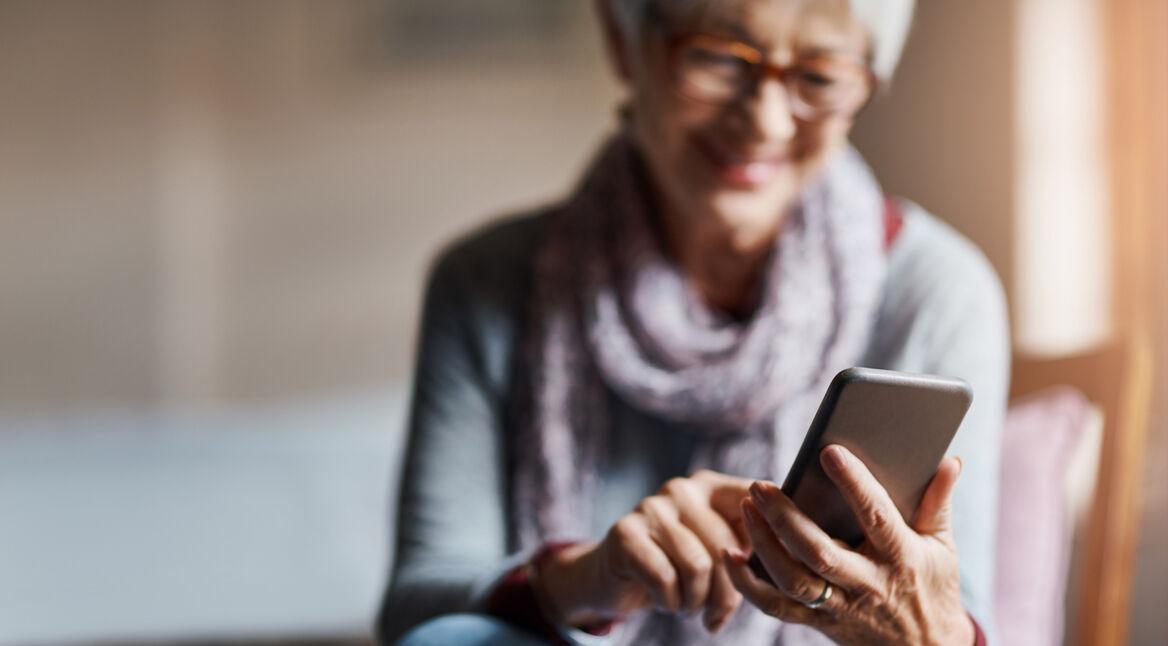 Auf dem Bild zum Thema Smartphones für Senior:innen ist eine ältere Frau mit grauen Haaren und Brille zu sehen, die entspannt auf einem Stuhl sitzt und lächelnd ein Smartphone bedient. Sie trägt einen Schal und wirkt zufrieden und konzentriert. Die Stimmung ist ruhig und positiv, das Licht ist warm und freundlich.