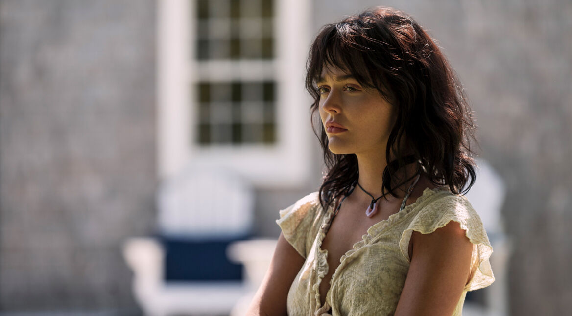 Auf dem Bild zur Serie Solange wir lügen Staffel 2 ist Emily Alyn Lind in ihrer Rolle als Cadence Sinclair Eastman zu sehen. Sie steht leicht seitlich im Bild, mit ernster Miene und nachdenklichem Blick. Ihr cremefarbenes Kleid mit Rüschen und das dunkle, leicht zerzauste Haar betonen den dramatischen Ton der Szene. Der Hintergrund zeigt unscharfe Gartenmöbel vor einer Hausfassade.