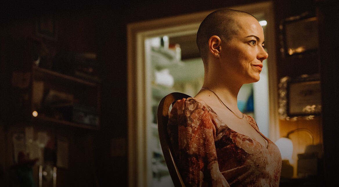 Emma Stone in Bugonia: Eine Frau mit kurz rasiertem Kopf sitzt in einem warm beleuchteten Raum und blickt entschlossen zur Seite.