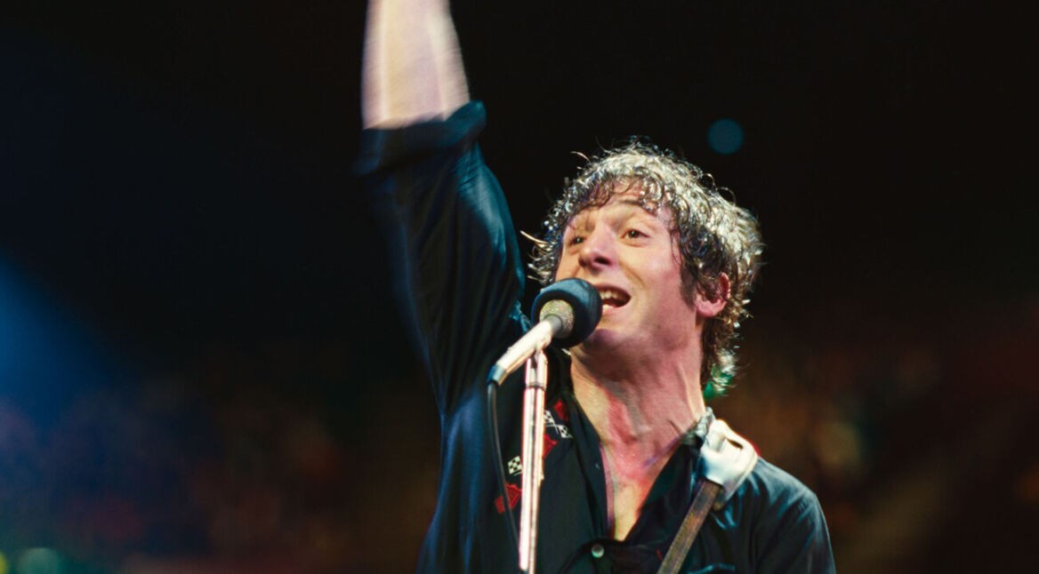 Jeremy Allen White wird in „Springsteen: Deliver Me From Nowhere”: Ein durchgeschwitzter Mann steht mit erhobenem Arm und Gitarrengurt über der Schulter auf einer Bühne vor einem Mikrofon.
