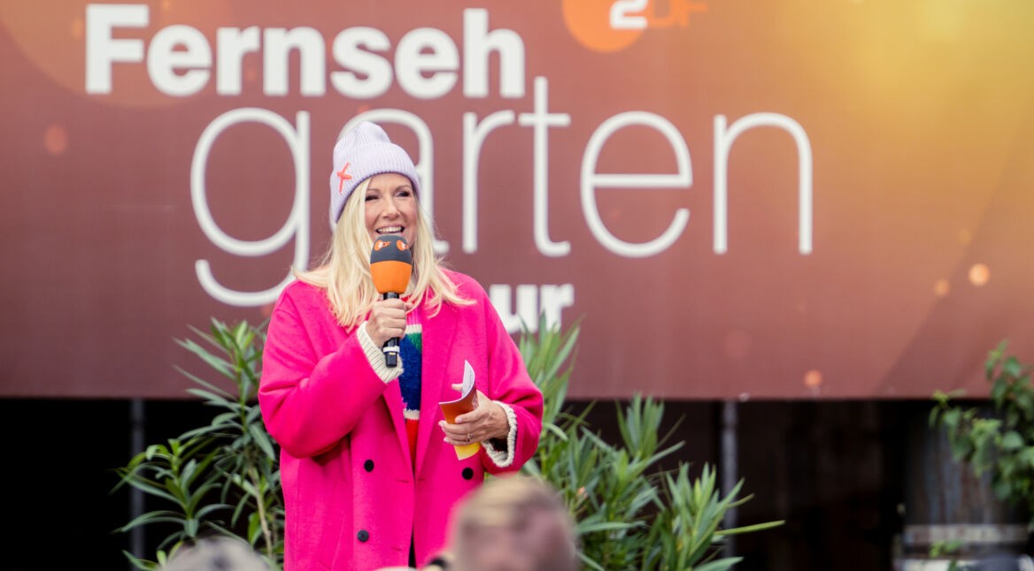 "ZDF-Fernsehgarten on tour": Andrea Kiewel steht in einem pinkfarbenen Mantel vor dem Sendungslogo. Sie hält ein Mikrofon und Moderationskarten in der Hand. Sie schaut lächelnd in Richtung Publikum.