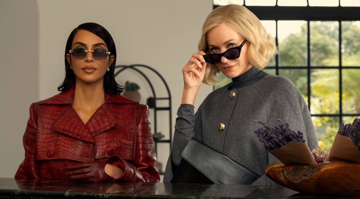In dieser stilvollen Szene aus All's Fair stehen Kim Kardashian als Allura Grant und Naomi Watts als Liberty Ronson nebeneinander. Allura trägt einen roten Mantel in Krokodiloptik mit passenden Handschuhen und Sonnenbrille, während Liberty in einem grauen Cape und mit hochgezogener Sonnenbrille selbstbewusst in die Kamera blickt. Die Stimmung ist kühl, kontrolliert und voller unterschwelliger Spannung.