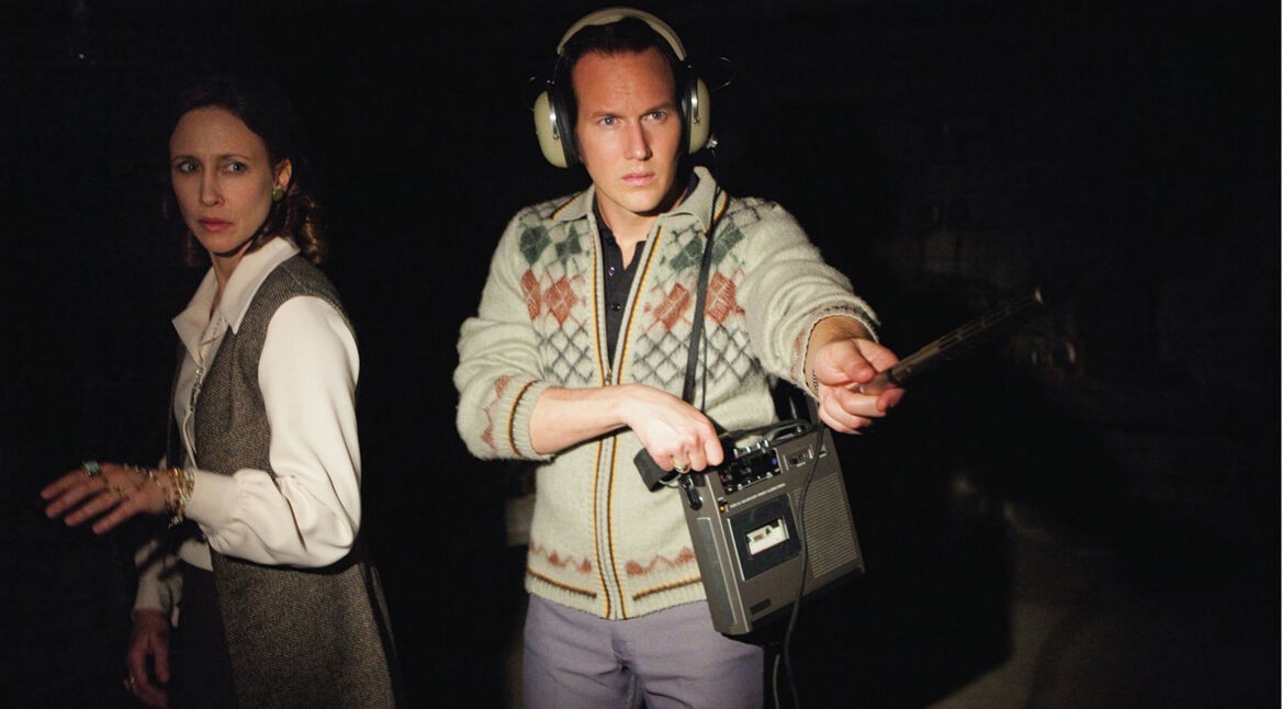Auf dem Bild aus dem Gruselfilm Conjuring – Die Heimsuchung sind Lorraine und Ed Warren zu sehen, gespielt von Vera Farmiga und Patrick Wilson. Ed trägt Kopfhörer und hält ein Tonbandgerät in der Hand, während er konzentriert in die Dunkelheit zeigt. Lorraine blickt ernst in eine andere Richtung. Die düstere Atmosphäre verstärkt die unheimliche Stimmung typischer Gruselfilme.