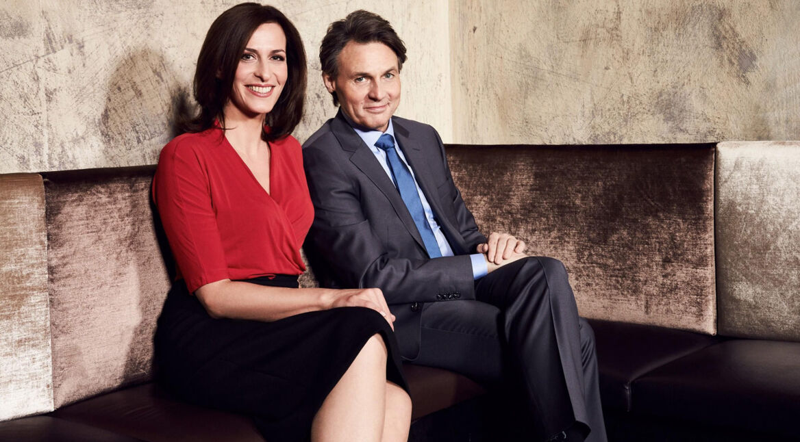 Auf dem Bild zur GZSZ-Besetzung sitzen Ulrike Frank und Wolfgang Bahro nebeneinander auf einem Sofa. Ulrike Frank trägt eine rote Bluse, Wolfgang Bahro einen grauen Anzug.