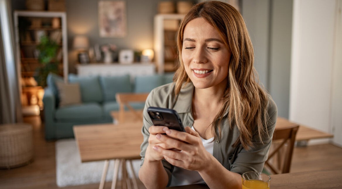 Im Symbolbild für den Handy Garantieverlängerung-Ratgeber sitzt eine lächelnde Frau entspannt in einem modern eingerichteten Wohnzimmer und schaut auf ihr Smartphone.