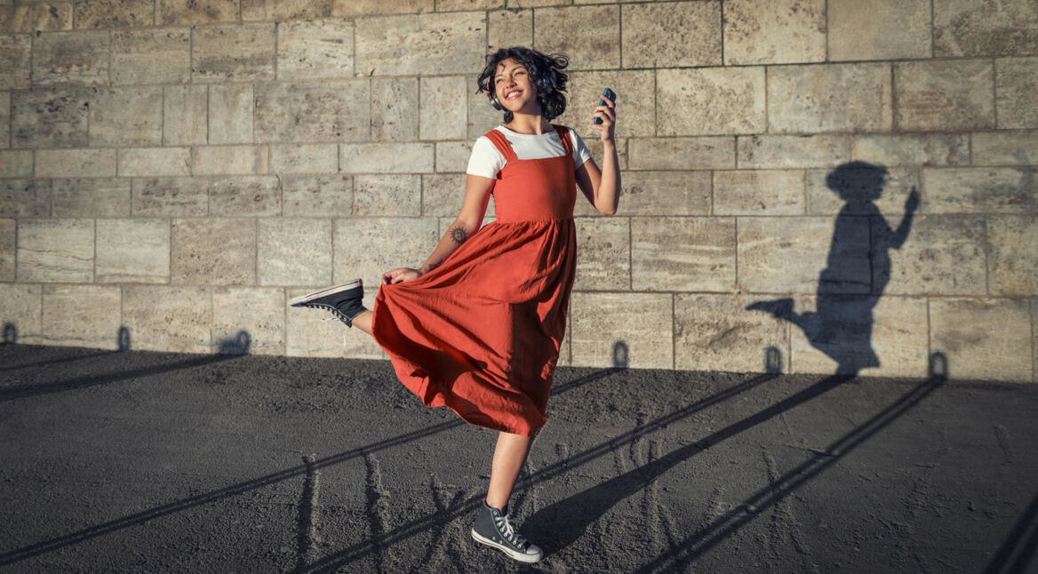 Datenverbrauch am iPhone: Junge Frau in rotem Kleid tanzt ausgelassen mit ihrem Smartphone in der Sonne vor einer Wand.