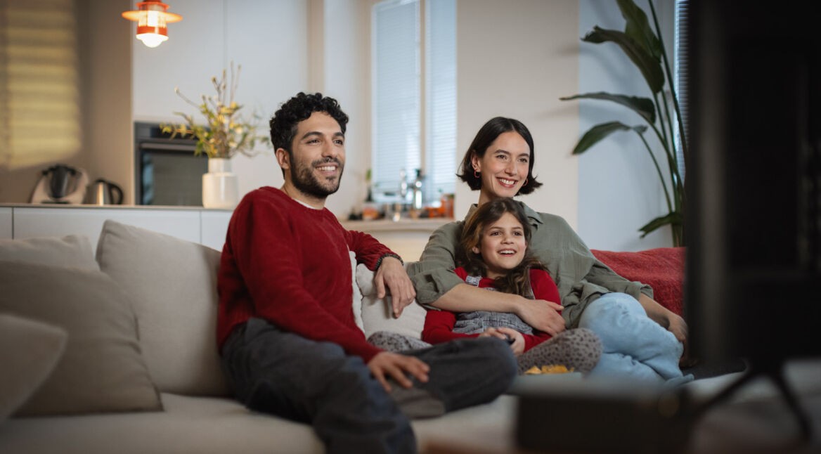Auf dem Bild zu den internationalen Sendern und TV-Paketen bei Vodafone sitzt eine fröhliche Familie gemeinsam auf dem Sofa und schaut entspannt Fernsehen. Ein Mann, eine Frau und ein Mädchen lachen und wirken völlig in das TV-Programm vertieft.