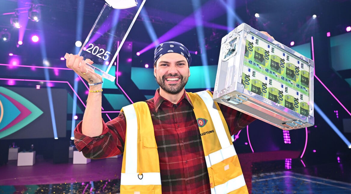 Der Sieger von „Promi Big Brother“ 2025 trägt ein kariertes Hemd, eine gelbe Weste und hält die Siegertrophäe sowie einen Geldkoffer hoch.