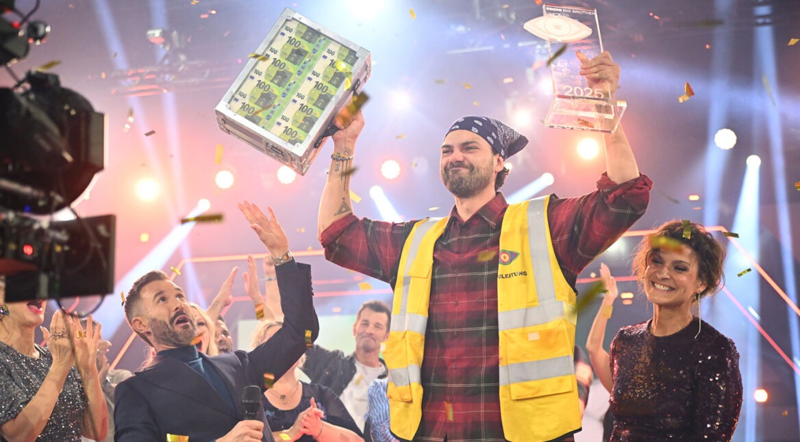 Auf dem Bild ist Jimi Blue Ochsenknecht als frisch gekürter Promi Big Brother Gewinner 2025 zu sehen. Er trägt eine gelbe Warnweste und hält stolz einen durchsichtigen Pokal sowie einen Koffer voller Geldscheine in die Höhe. Um ihn herum jubelt das Publikum, Konfetti fliegt durch die Luft, die Stimmung ist ausgelassen und feierlich.