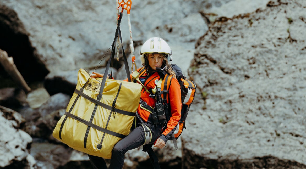 Katharina Strasser (Luise Bähr) hängt vor einer Felswand an einem Rettungstau. Ebenfalls am Tau ist ein gelber zusammengelegter Bergesack befestigt.