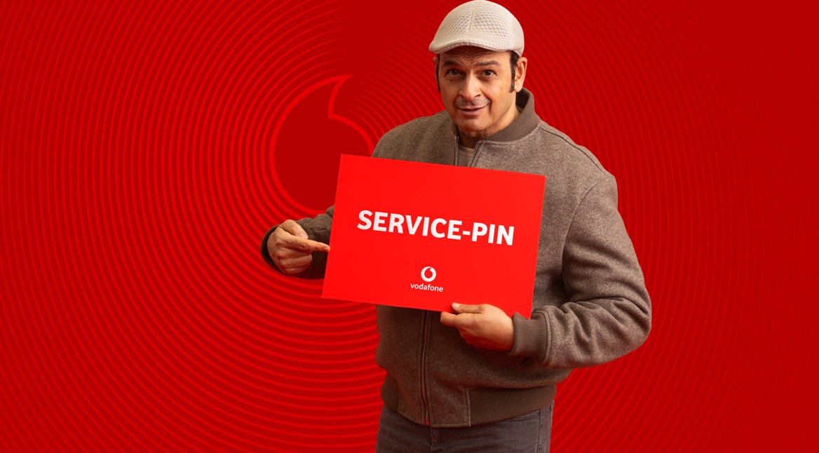 Kaya Yanar ist auf dem Bild zur Vodafone Service-PIN Kampagne zu sehen, wie er ein rotes Schild mit der Aufschrift „SERVICE-PIN“ hält und darauf zeigt.