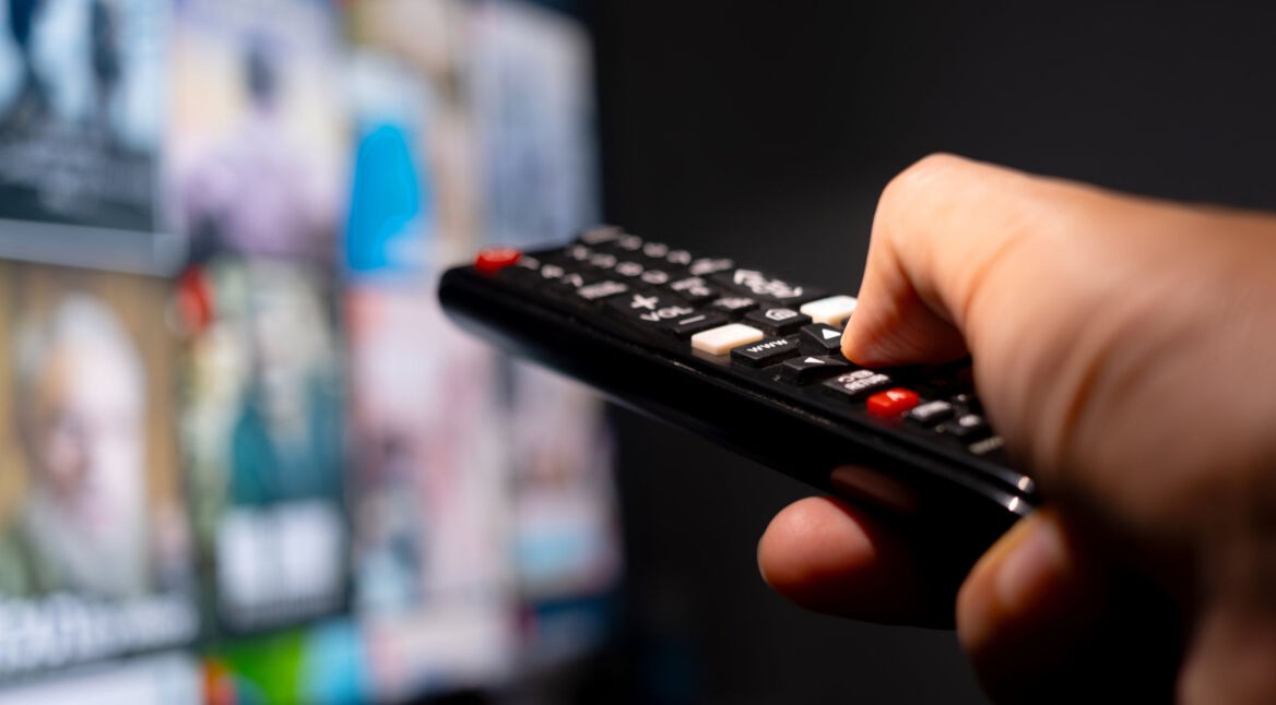 Eine Hand hält eine Fernbedienung und zeigt auf einen TV, der im verschwommen im Hintergrund zu sehen ist.