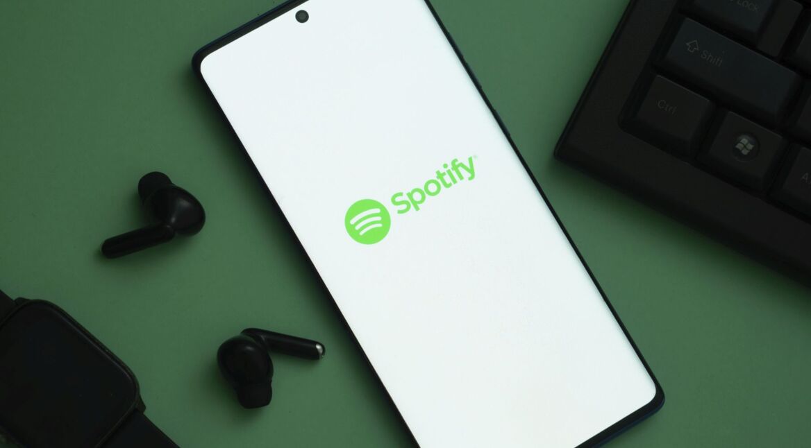 Das Logo von Spotify ist auf einem Handy zu sehen, links davon liegen schwarze Kopfhörer.