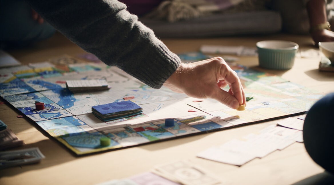 Auf dem Bild zu Brettspiel-Filme ist eine Szene am Spieltisch zu sehen, bei der eine Hand eine gelbe Spielfigur auf einem bunt gestalteten Spielbrett platziert. Rund um das Brett liegen Karten, Spielgeld und Spielsteine. Die Stimmung wirkt entspannt und familiär, Tageslicht fällt sanft auf die Szene.