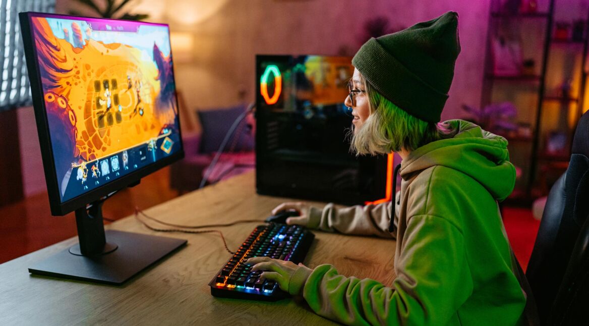 Beim Cloud-Gaming Vergleich zeigt dieses Bild eine junge Person mit grüner Mütze und Kapuzenpullover, die an einem RGB-beleuchteten Gaming-Setup spielt, während ein farbenfrohes Spiel auf dem Monitor läuft.