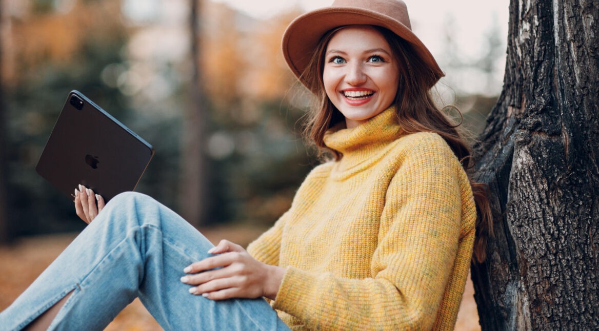 Ein junge Frau lehnt an einem Baum. Sie hält ein iPad in der Hand und lächelt in die Kamera.