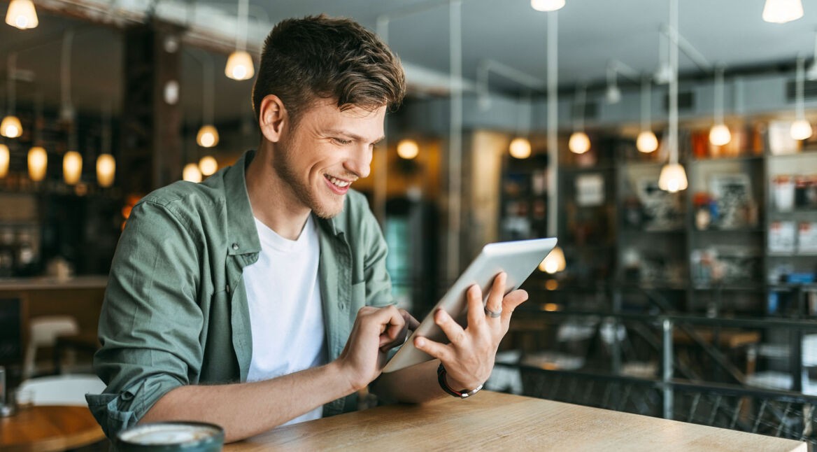 Ein junger Mann sitzt in einer Buchhandlung und schaut auf ein Tablet.