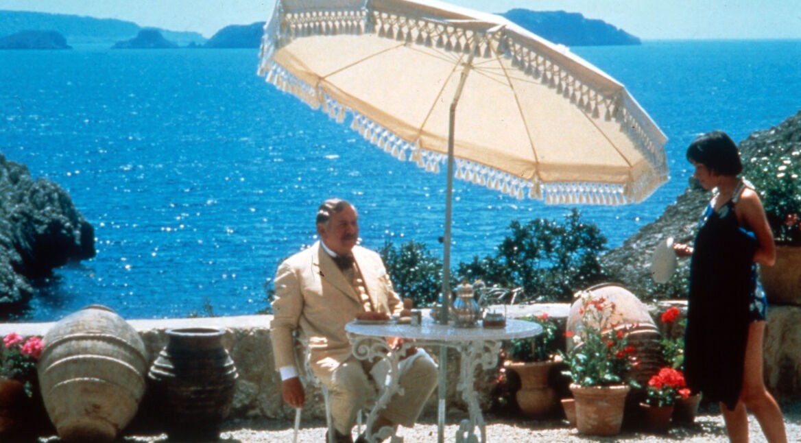 Filmszene aus "Das Böse unter der Sonne". Peter Ustinov, als Meisterdetektiv Hercule Poirot, sitzt auf einer sonnigen Terrasse an einem Tisch. Im Hintergrund ist das Meer zu sehen.