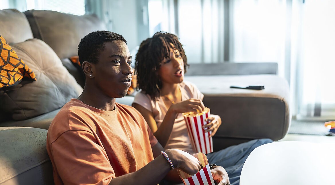 Ein junges Paar sitzt vor dem Sofa auf dem Boden und schaut Fernsehen.