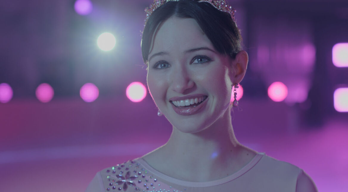 Madelyn Keys in Finding Her Edge: Eine junge Eiskunstläuferin mit Diadem lächelt im Schein bunter Lichter in der Eishalle.