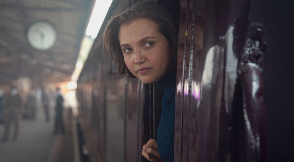 Mia McKenna-Bruce als Bundle schaut in einer Szene von "Agatha Christie's Seven Dials" aus einem Zugfenster heraus.
