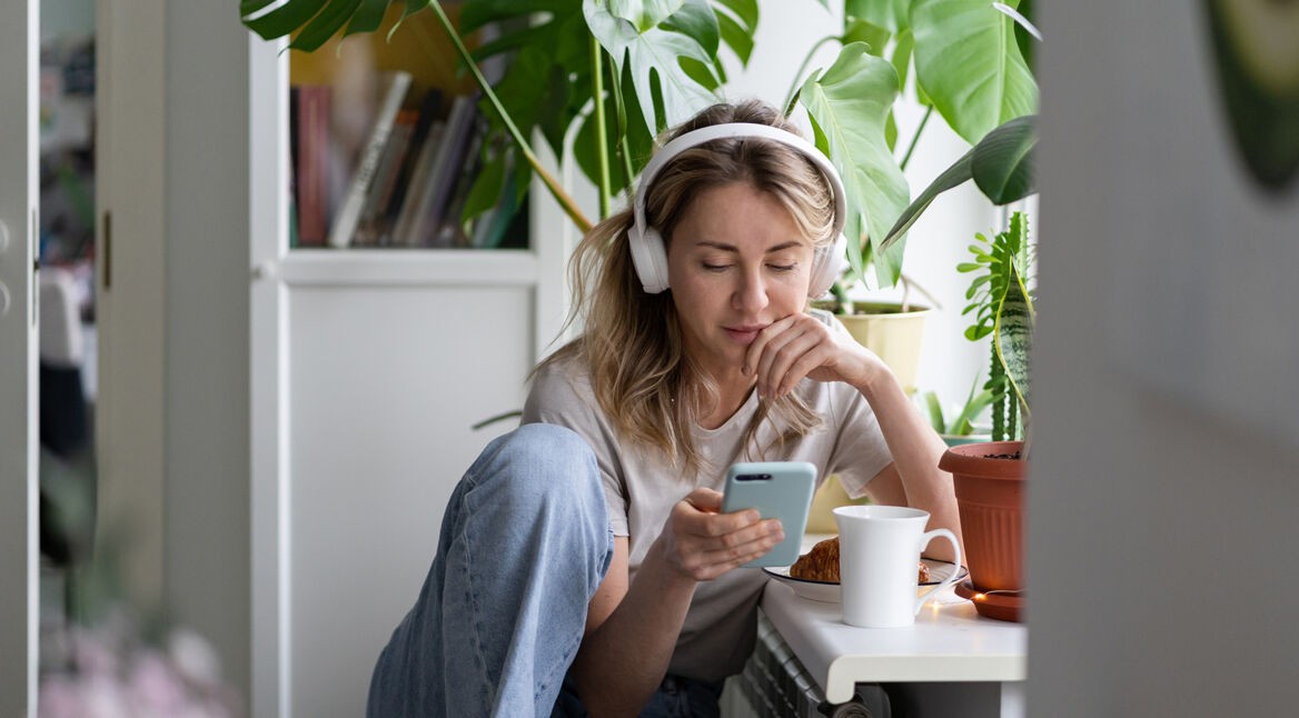 Eine junge Frau sitzt entspannt an einem Tisch im Wohnzimmer, umgeben von Zimmerpflanzen. Sie trägt weiße Kopfhörer, schaut auf ihr Smartphone und hört offenbar konzentriert einen Beitrag. Vor ihr stehen ein Croissant und eine weiße Tasse. Die Szene vermittelt Ruhe und Fokus – passend zum Thema beste ARD-Podcasts.