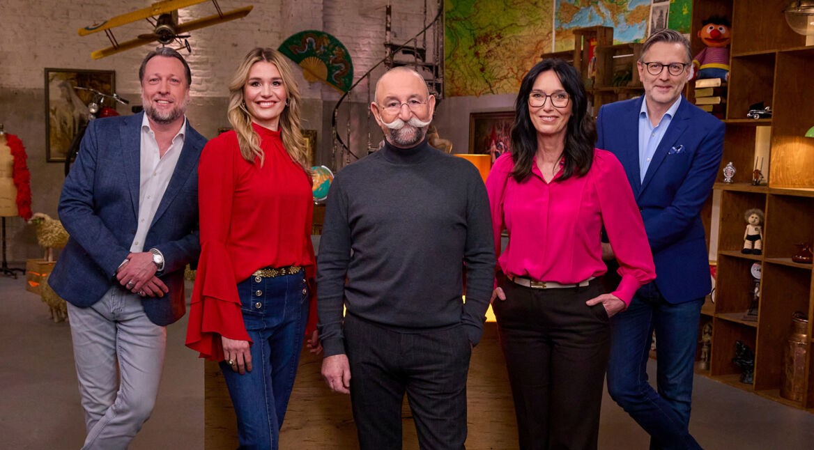 Auf dem Bild mit den Bares für Rares-Expert:innen stehen fünf bekannte Gesichter der Sendung lächelnd nebeneinander im Studio. Von links nach rechts sind Patrick Lessmann, Bianca Berding in roter Bluse, Moderator Horst Lichter mit markantem Schnurrbart, Heide Rezepa-Zabel in pinker Bluse und Detlev Kümmel im blauen Sakko zu sehen. Die Stimmung wirkt freundlich und professionell.