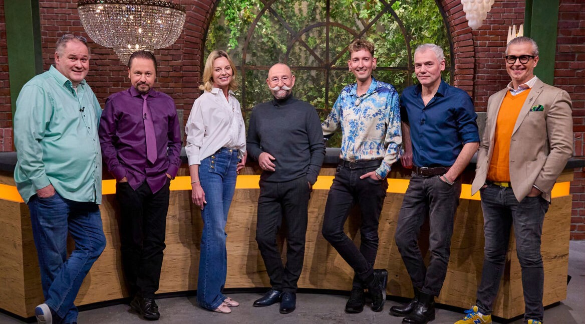 Auf dem Bild sind die Bares für Rares Händler Walter Lehnertz, Wolfgang Pauritsch, Susanne Steiger, Fabian Kahl, Christian Velten und Anaisio Guedes gemeinsam mit Moderator Horst Lichter zu sehen. Sie stehen gut gelaunt im Studio der ZDF-Sendung, vor einer Kulisse mit rustikaler Backsteinwand und großen Fenstern mit Blick ins Grüne.