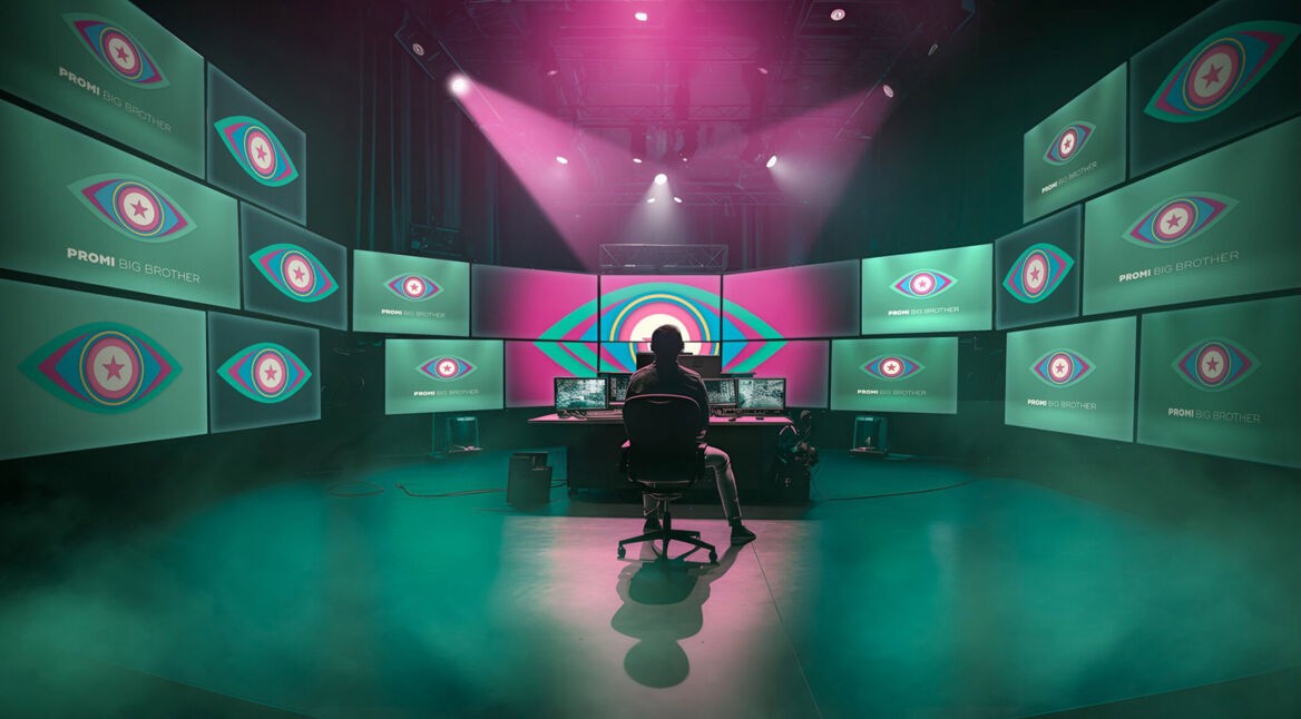 In diesem Bild zur Show Promi Big Brother sitzt eine Person in einem abgedunkelten Regieraum vor einer riesigen Monitorwand. Auf allen Bildschirmen ist das ikonische Auge-Logo von Promi Big Brother zu sehen. Die Szene ist in grünliches Licht und Nebel getaucht, darüber strahlen dramatische pinke Scheinwerfer – die Stimmung wirkt geheimnisvoll und überwachend.