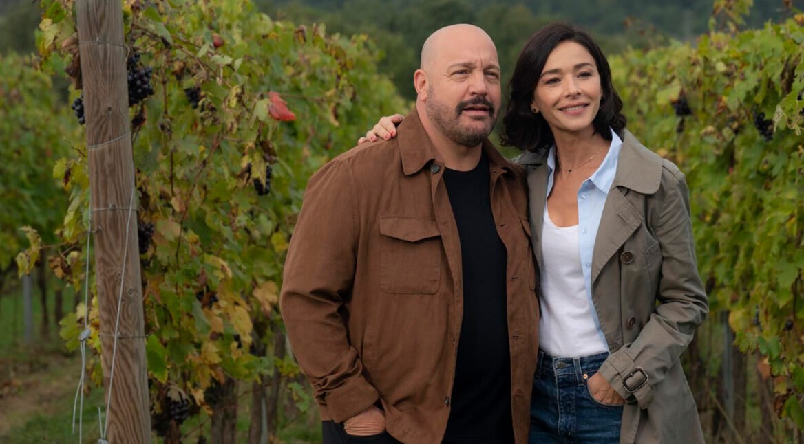 Auf dem Bild zum Film Solo Mio mit Kevin James stehen Matt, gespielt von Kevin James, und Gia, gespielt von Nicole Grimaudo, eng beieinander inmitten eines Weinbergs. Beide wirken gelöst und glücklich – Matt trägt eine braune Jacke, Gia einen beigen Mantel. Die herbstlich gefärbten Reben im Hintergrund erzeugen eine ruhige und romantische Stimmung.