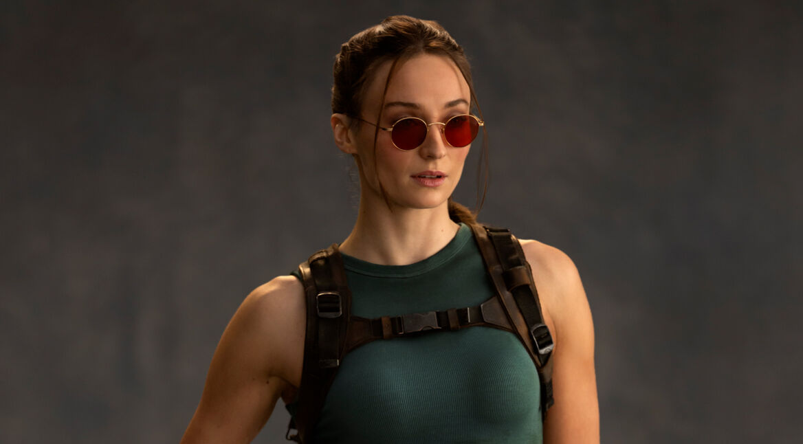 Auf dem Bild zur Tomb Raider Serie ist Sophie Turner als Lara Croft zu sehen. Sie trägt ein enges, dunkelgrünes Tanktop und ein taktisches Geschirr mit Schnallen. Ihre Haare sind zurückgebunden, einige Strähnen fallen locker ins Gesicht. Auffällig sind die runden, roten Sonnenbrillen, die ihrem entschlossenen Gesichtsausdruck eine coole Note verleihen.