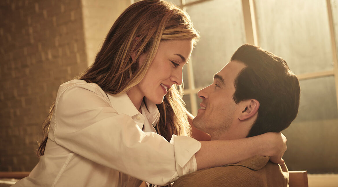 Sarah Pidgeon sitzt als Carolyn Besette auf dem Schoß von Paul Anthony Kelly als John F. Kennedy Jr. Sie hat ihre Arme um seinen Hals gelegt und die beiden schauen sich tief in die Augen.